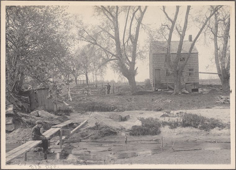 Black-and-white photograph from 1902 showing a small house in Mill Neck, Long Island, set behind a shallow stream with a narrow wooden footbridge in the foreground and a person seated at the edge of the bridge, surrounded by trees and a fenced field.